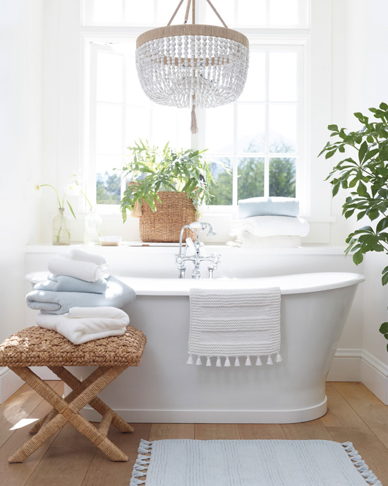 Light and airy bathroom, white free-standing tub, big window with light streaming in, house plants and featuring bath mats and towels.