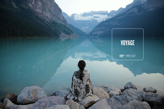 Girl sitting on rocks, wrapped in a warm blanket, looking out over mountains and a still crystal blue body of water. 