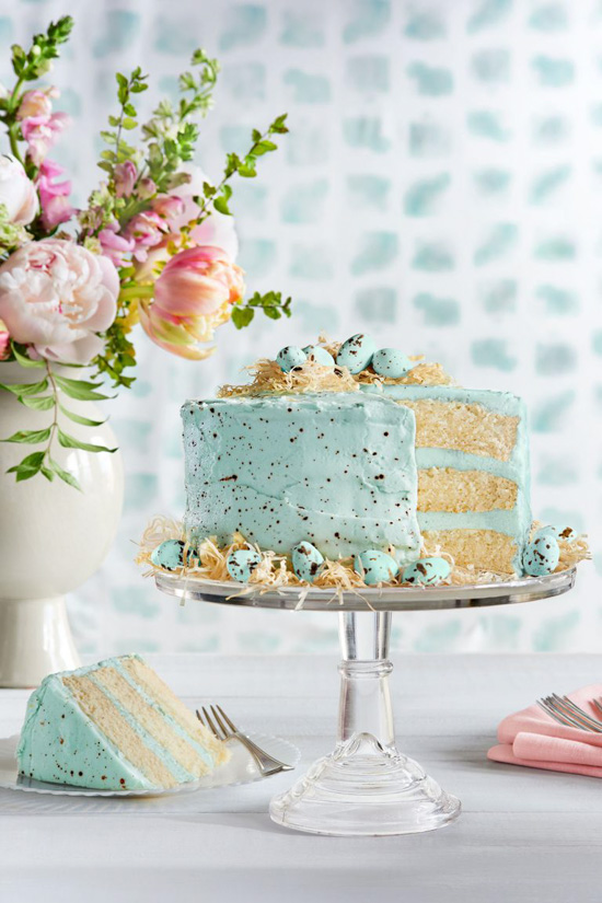Bouquet of spring flowers, a glass cake stand topped with a three layered cake covered in a speckled robin egg blue frosting with a slice cut out and set to the side.