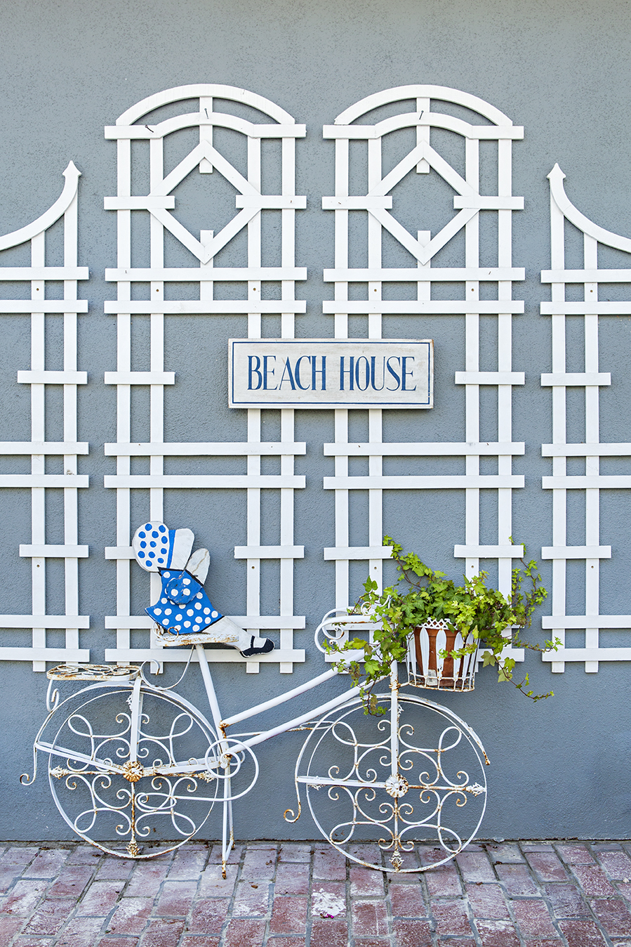 A blue exterior wall juxtaposed to a brick sidewalk with a rod iron decorative bicycle leaned against a white lattice design.