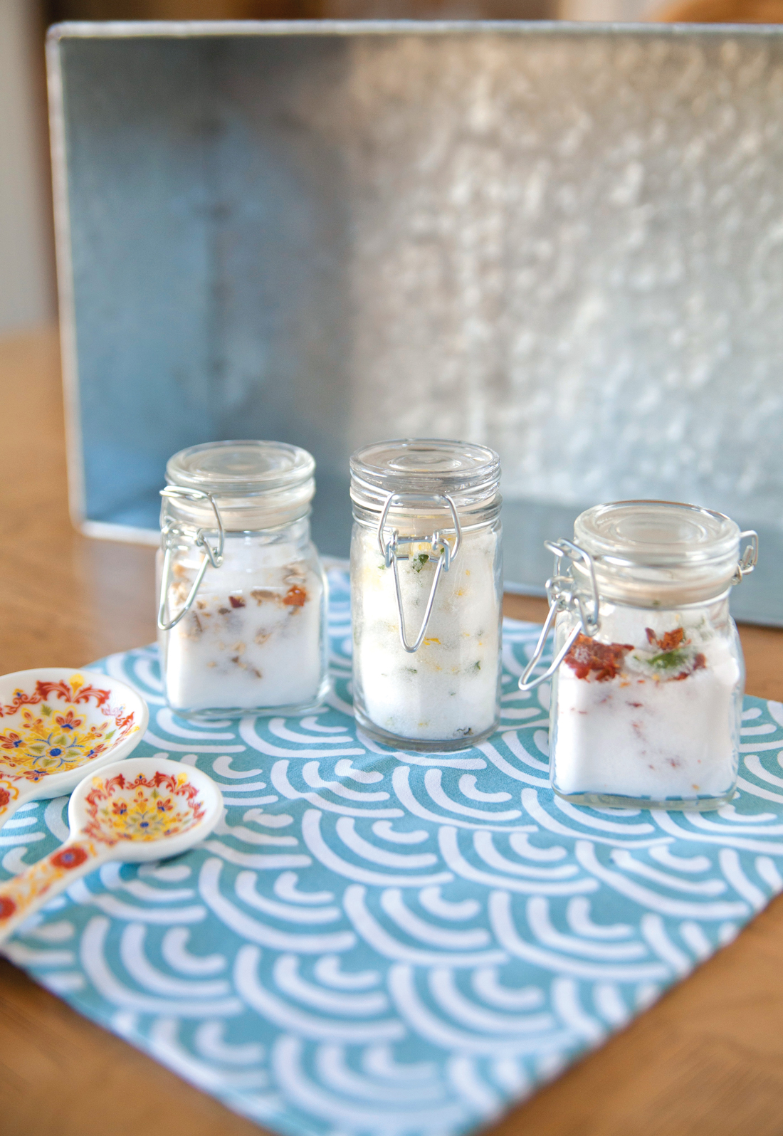Three small glass canisters filled with custom seasoned salts on a linen napkin with a silver serving tray in the background. 
