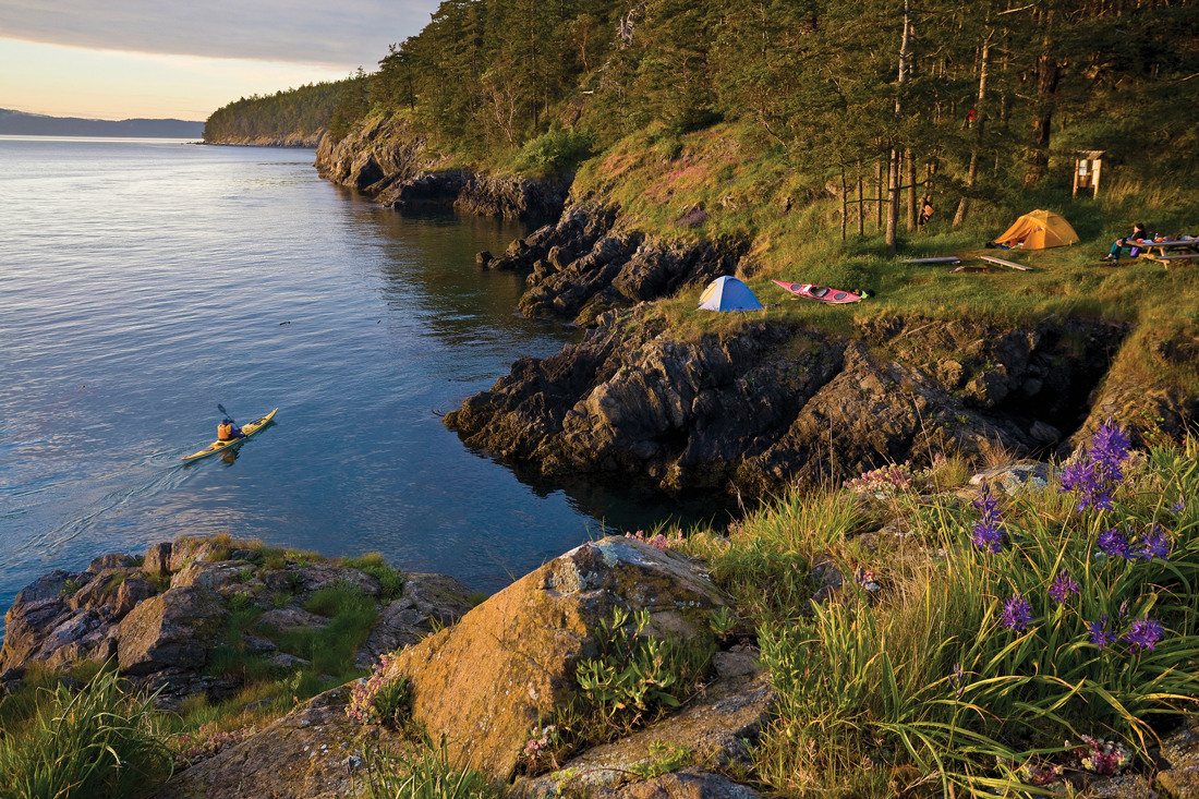 Look out over cliffs leading to the ocean, tents set up on the flatlands and a kayaker in the water. 