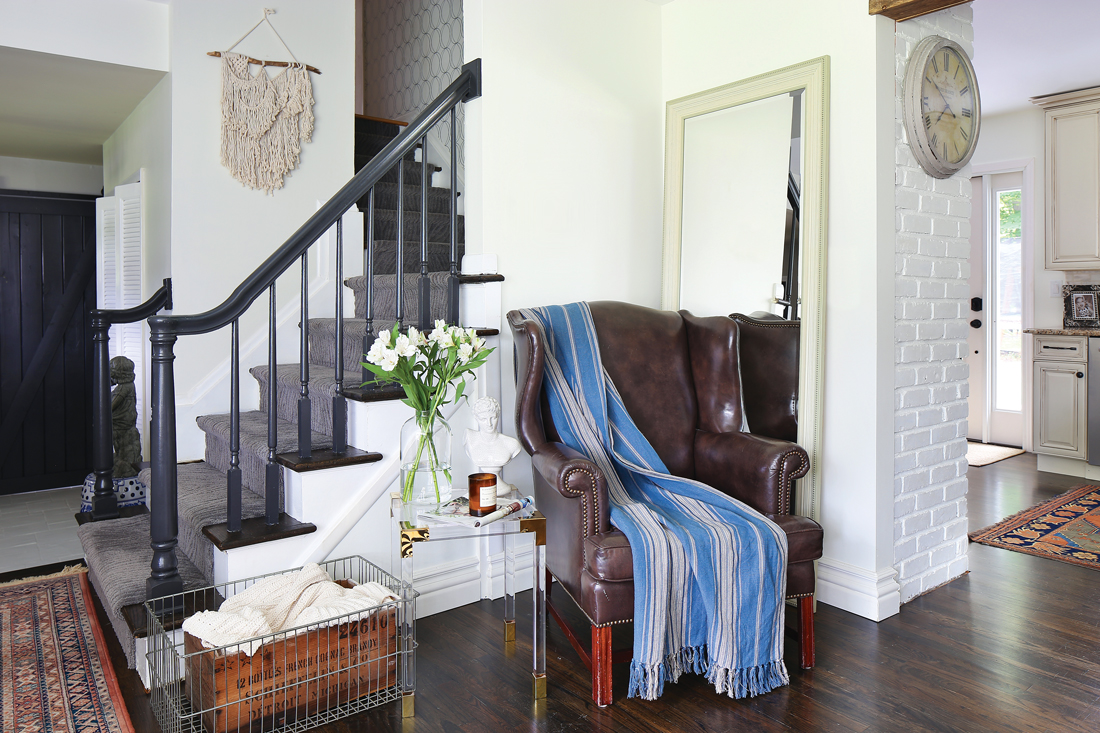 Dark railed stairs frame out a sitting nook with a clear and gold modern endurable and a vintage leather wingback chair. 