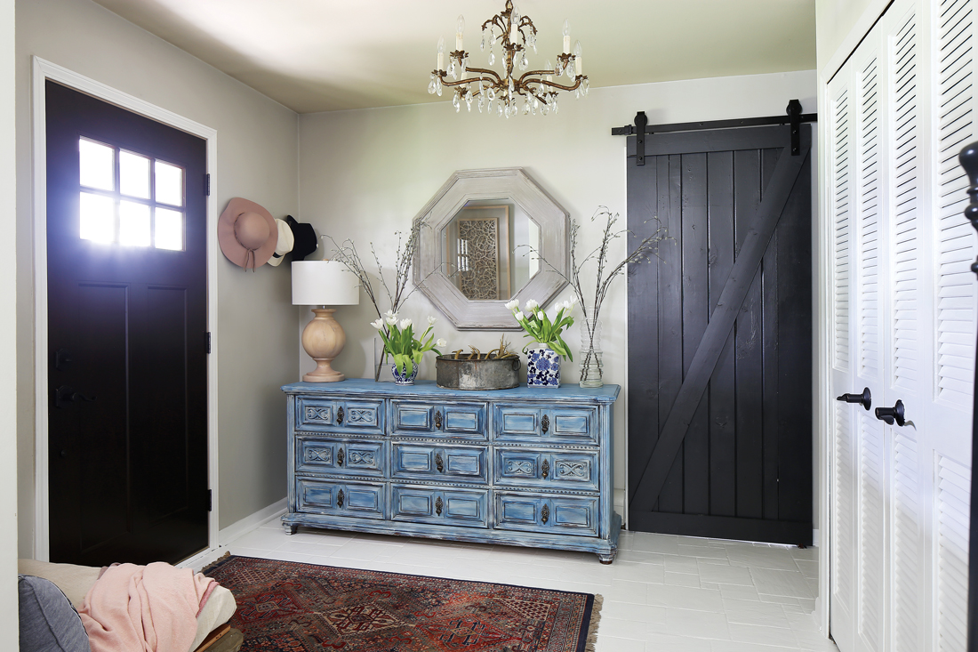 View from the interior of the home at the entryway. The foyer includes a Persian runner, sliding barn door, and an octagonal shaped mirror hanging over a vintage distressed dresser. 