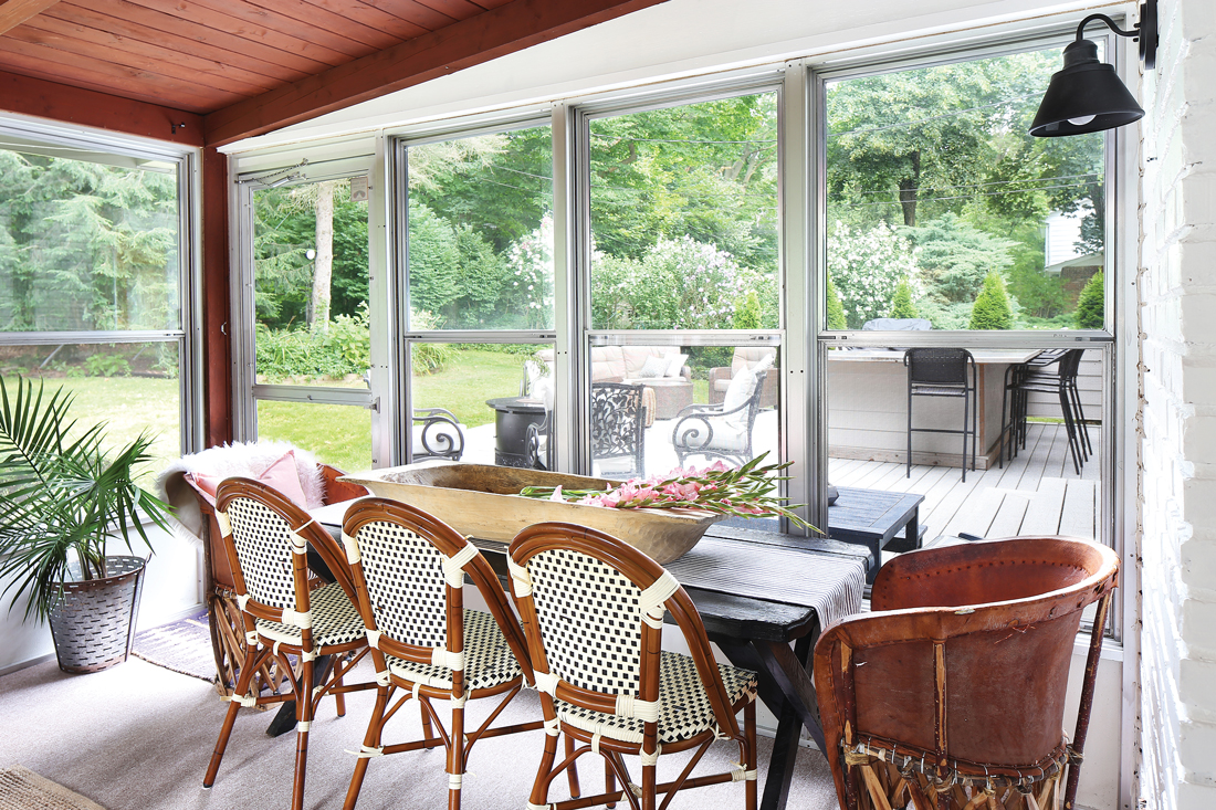 Enclosed patio with full dining space. Black and white whicker bistro chairs mixed with Mexican leather chairs and a wall of windows looking out into the yard. 