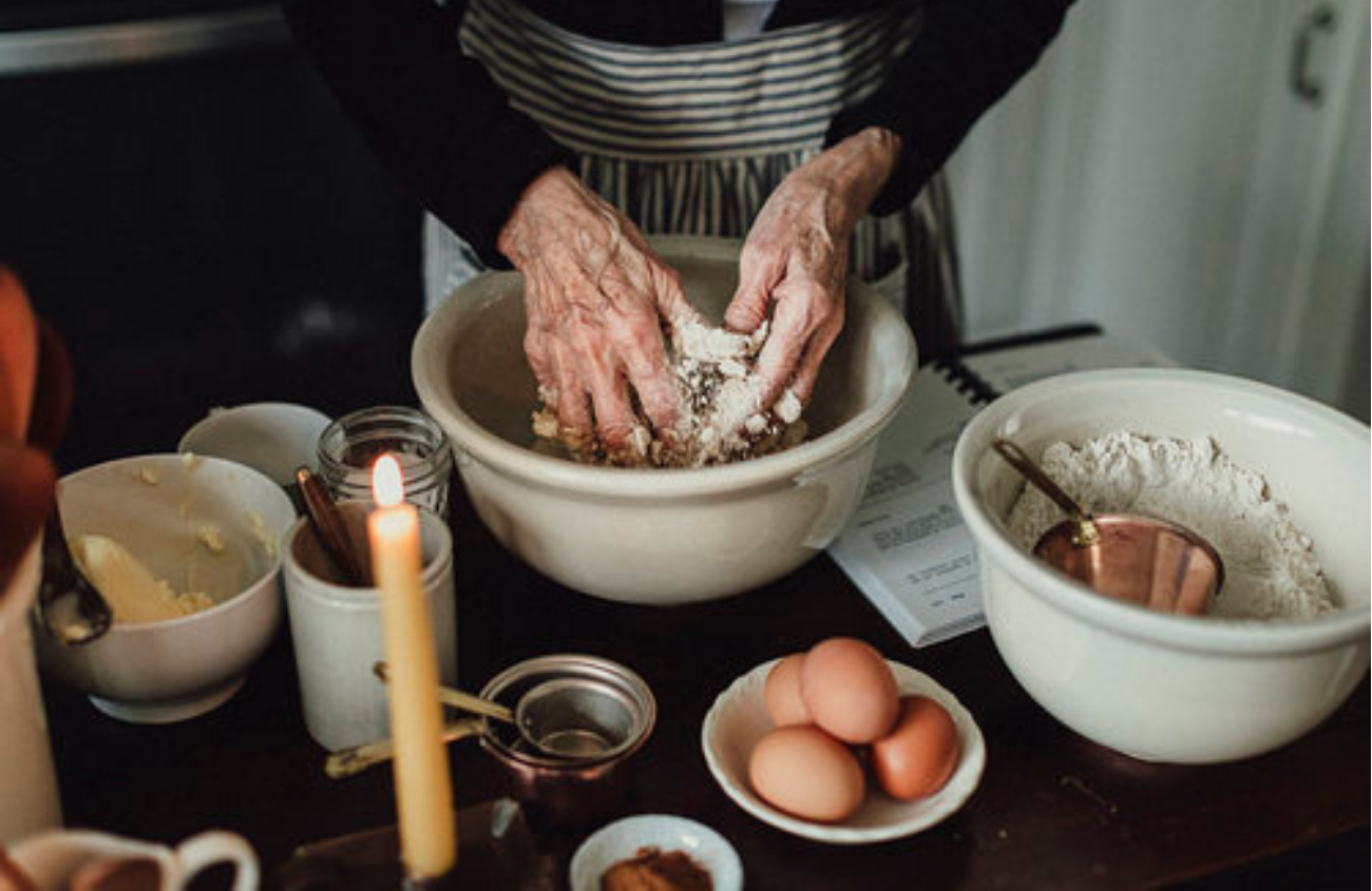 A cluttered table covered in baking supplies and an old woman's hands kneading dough.