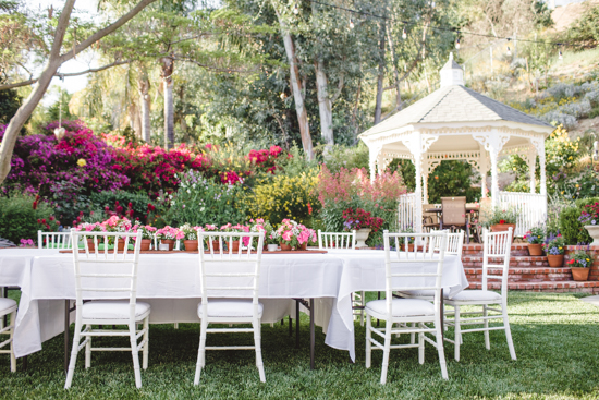 Outdoor table with white table cloths and chairs with a white gazebo in the background. 