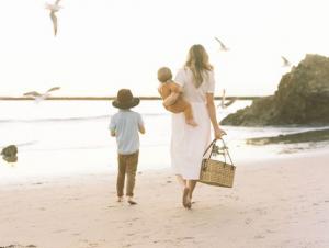 Mother walking with son down the beach during sunset with a baby on one hip and a picnic basket in the other.
