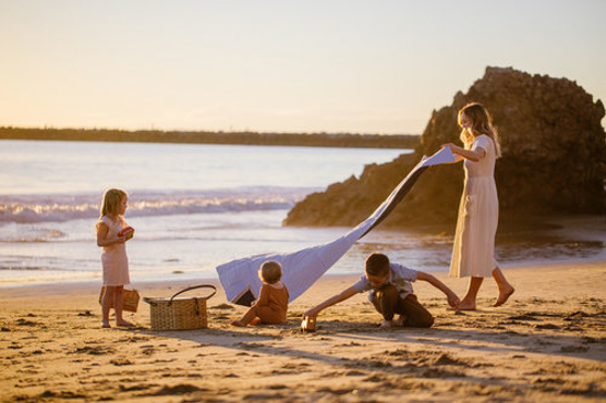 Mother and her three little ones on the beach at sunset laying out a blanket on the sand for a picnic. 