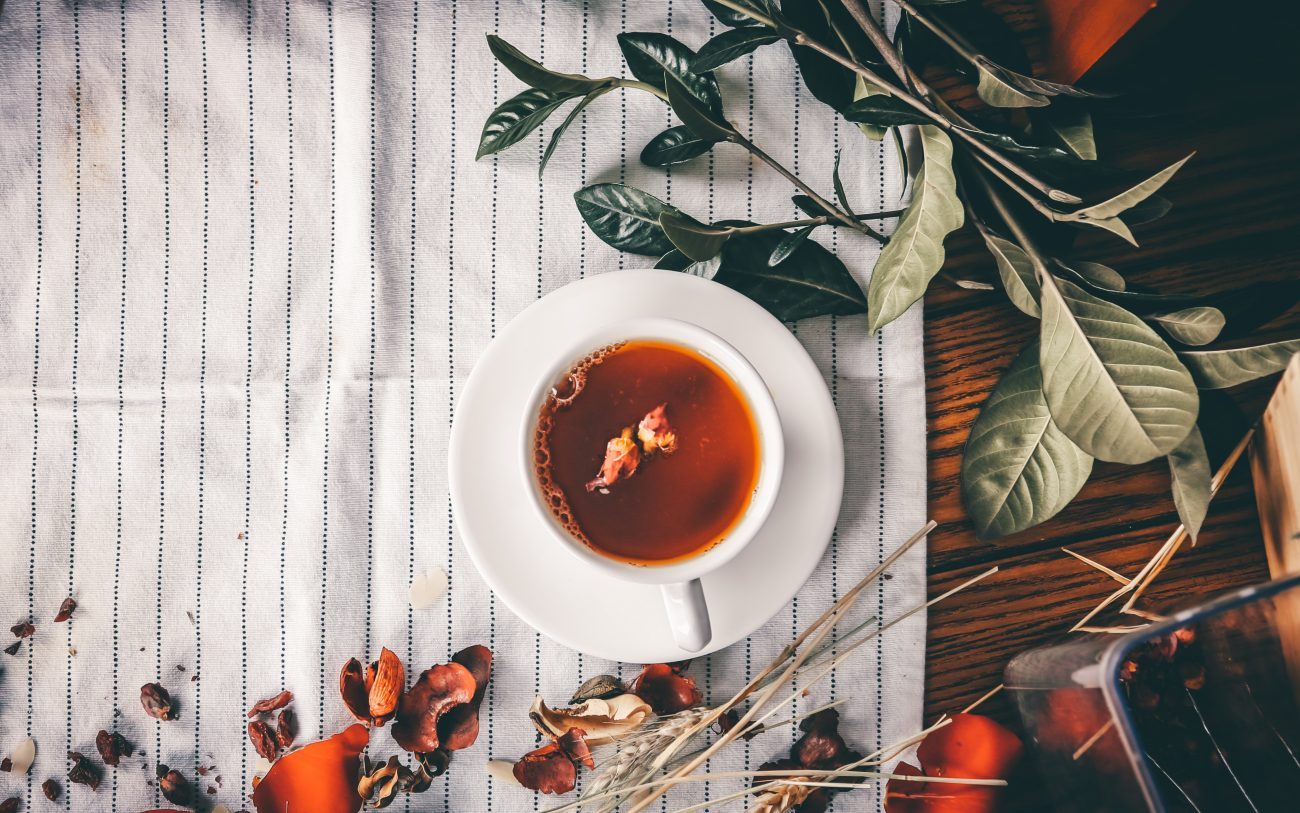 An aerial shot of a cup of black tea in a tea cup laid on a dish towel and surrounded by herbs and shrubbery.