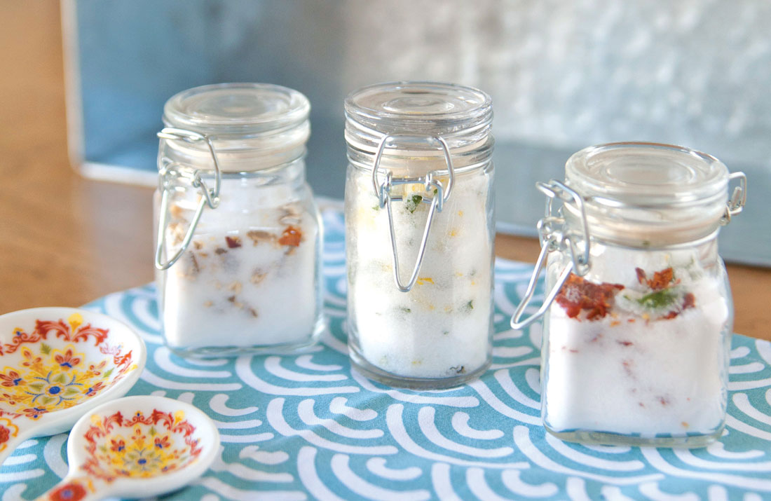 Three small glass canisters filled with custom seasoned salts on a linen napkin with a silver serving tray in the background.