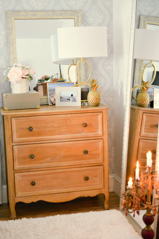 Wooden dresser topped with gold accented frames, romantic floral arrangement and a large mirror and desk lamp. 