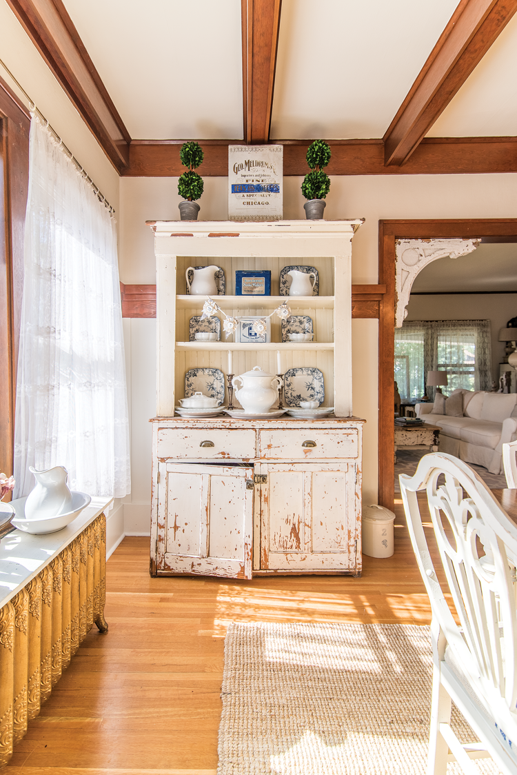 A white rustic, distressed hutch near an original radiator with an exposed wooden ceiling beams. 