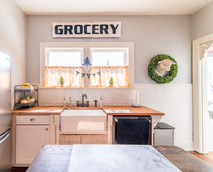 Butcher block countertops and a bright white backdrop set the kitchen scene with an antique "grocery" sign mounted above the farmhouse sink.