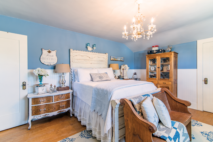 Charming farmhouse inspired bedroom with white and blue accents, a crystal chandelier, and a restored wooden church pew at the foot of the bed.