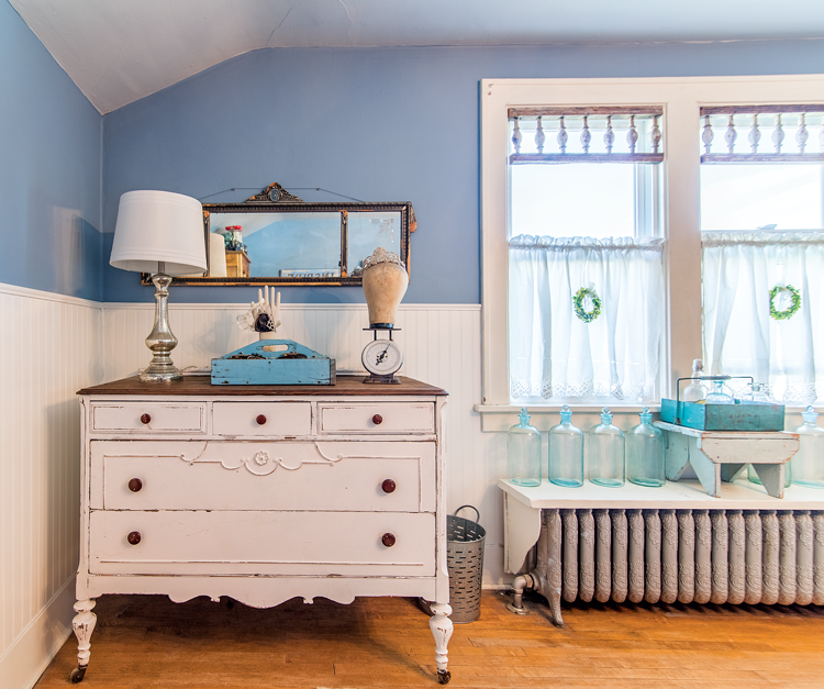 A found antique mirror hanging above a vintage detailed white dresser near bright craftsman style windows. 