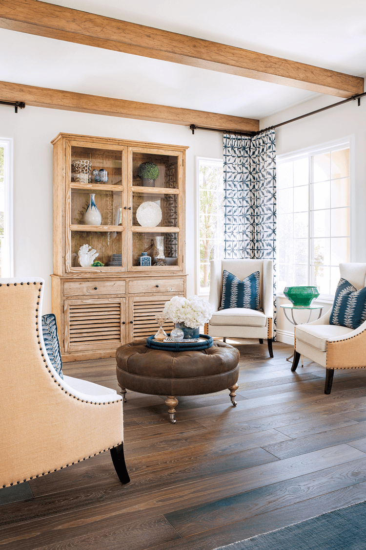 Peek into this sitting room with exposed ceiling beams, cream colored armchairs around a leather, tufted coffee table and shibori curtains framing the windows. 