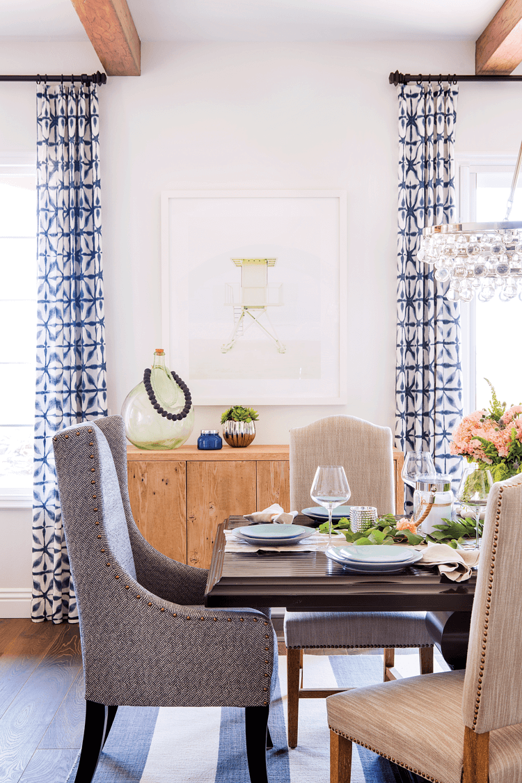 A substantial dark brown dining table surrounded by upholstered dining chairs. Above are exposed beams and shibori curtains framing the windows in the background. 