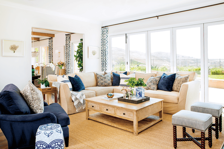 Living room with a full wall of fold-away windows, filled with a large sand colored sectional, wooden coffee table and navy blue armchair. 