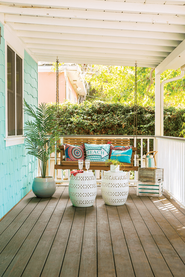 Wooden slatted front porch with white railing and a swinging wooden bench swing with colorful beach inspired pillows and accents. 