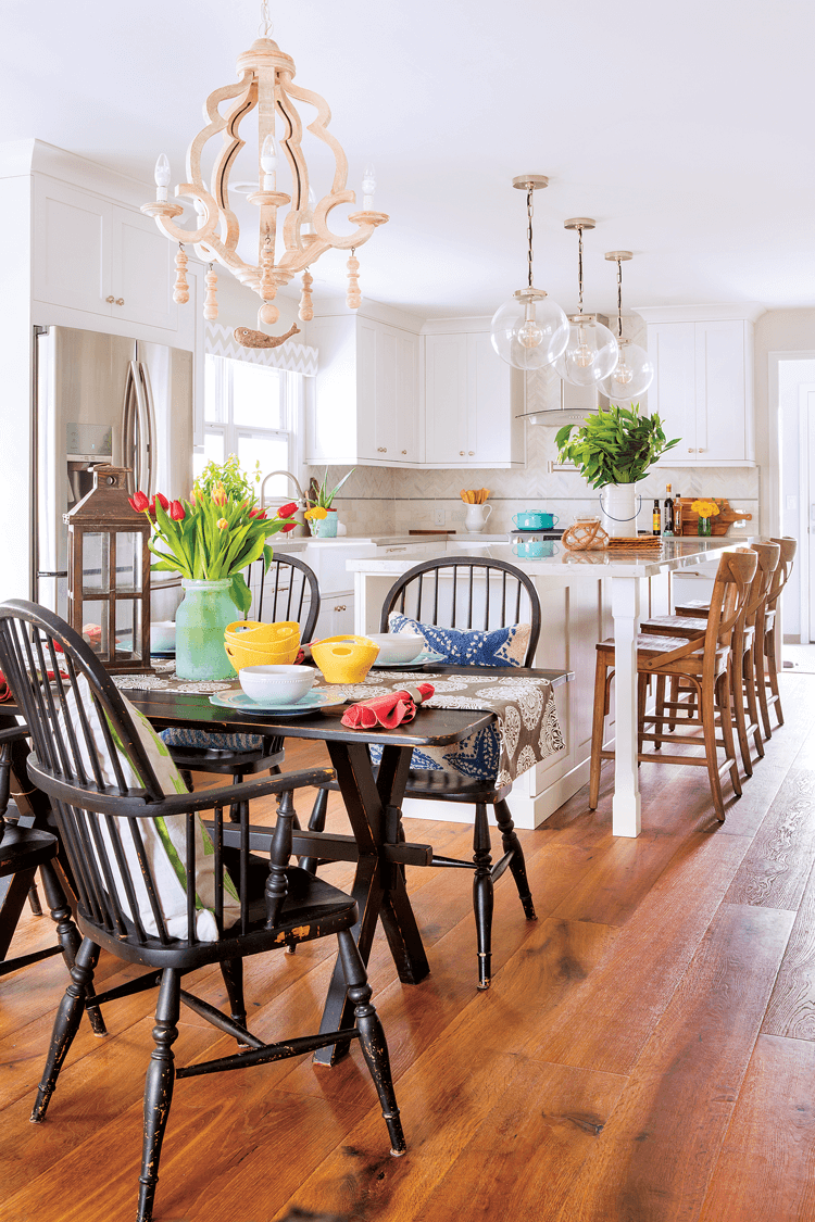 Distressed dark wooden farmhouse chairs and dining table covered in bright and colorful tulips. Looking into an open concept white and bright colorful beach cottage kitchen. 