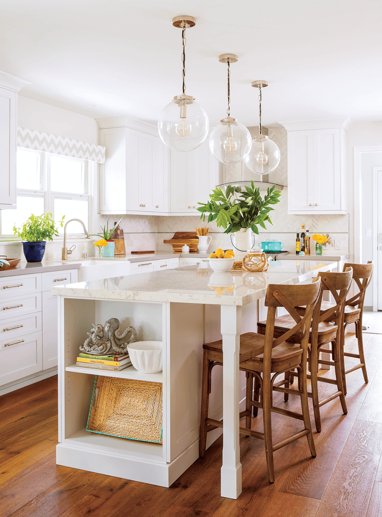 Marble topped large kitchen island in the center of a white and bright kitchen accented by globe pendant lights and fresh greenery. 