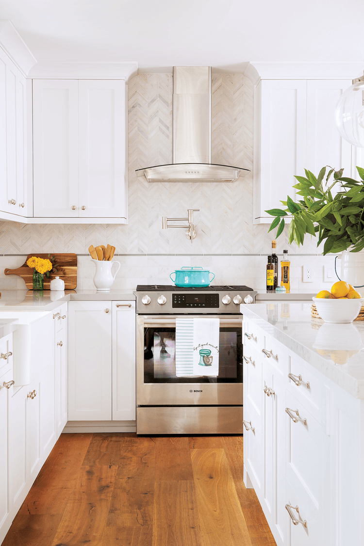 Looking down the galley of the kitchen toward the sleek stainless steel range and hood displayed in front of the marbled chevron backsplash. 