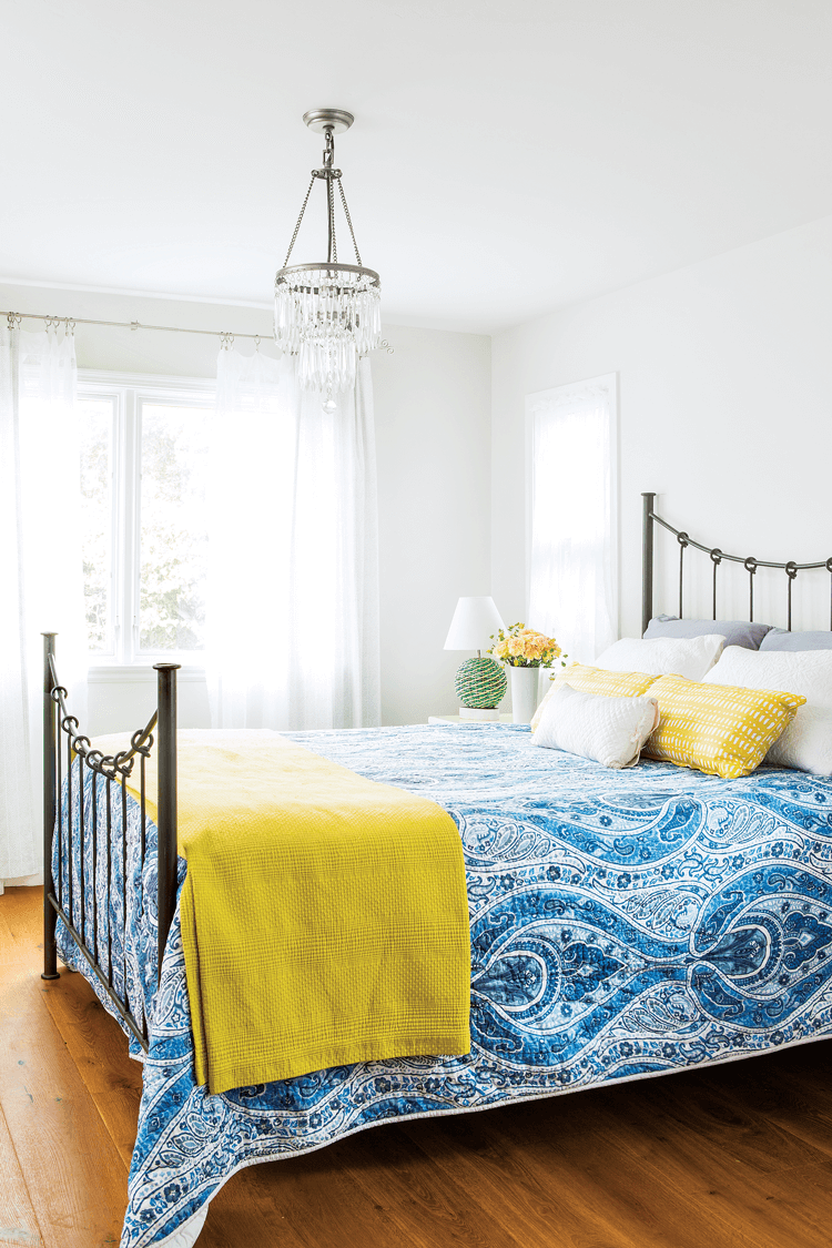 Bright and colorful beach cottage bedroom, white walls with a crystal chandelier and a bright blue quilt on the rod-iron bed. 