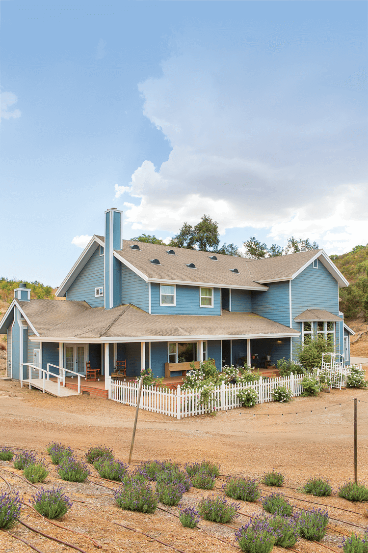 A custom designed farmhouse painted a shade of eggshell blue surrounded by a lovely garden and a white picket fence. 