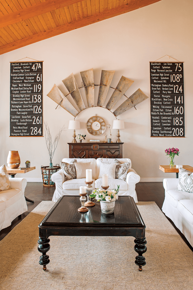 Vaulted exposed wood ceilings in an all white room with covered sofas. Large windmill pieces mounted on the wall and a bold, black farmhouse coffee table. 