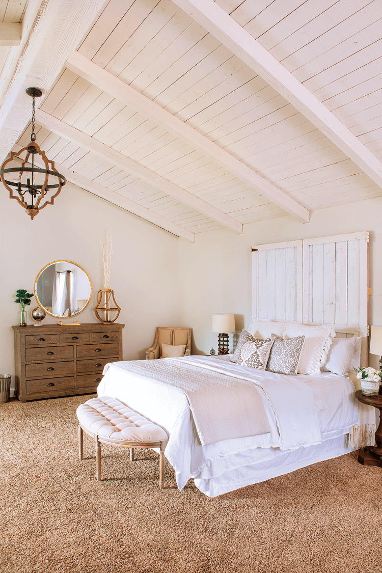 An all white, rustic style bedroom complete with exposed, vaulted ceilings and warm wooden accents like the dresser and chandelier.