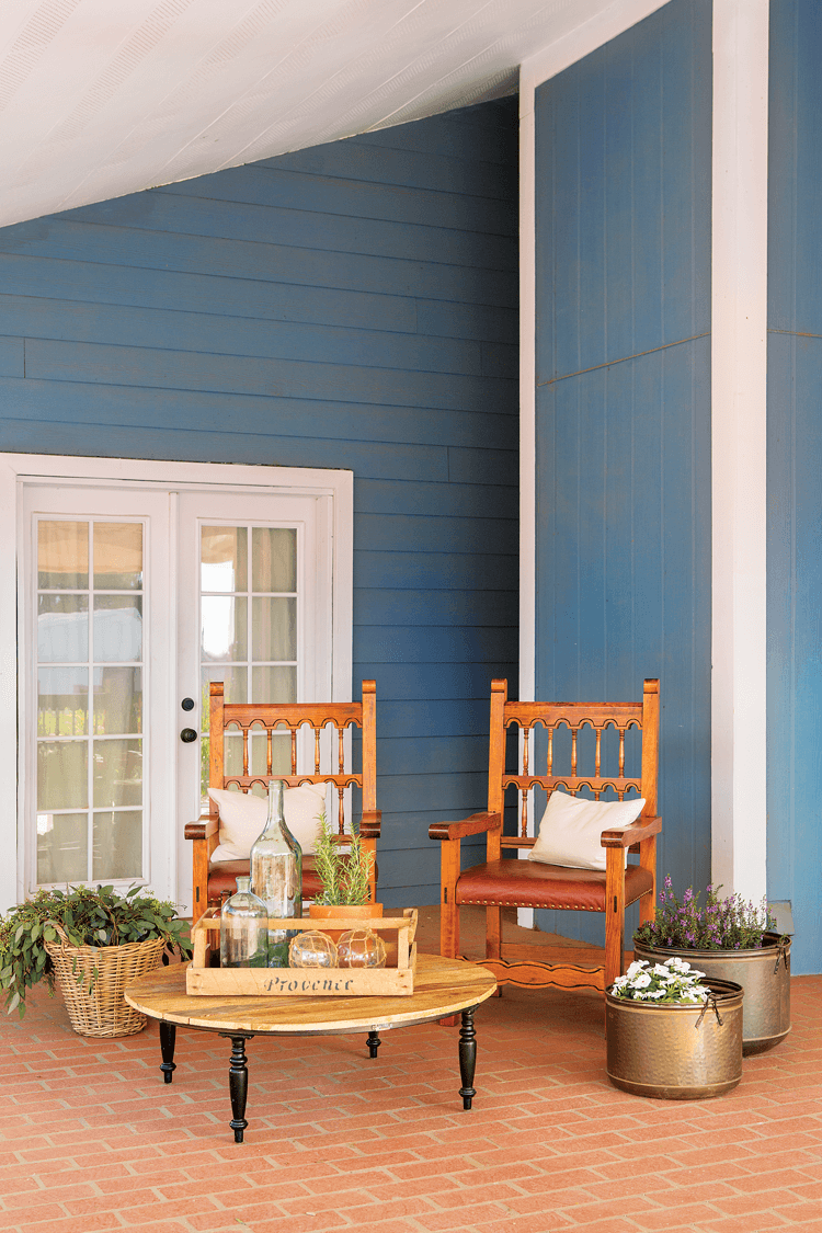 Beautiful French doors leading into the house from the porch. Ornate wooden chairs for a small seating area surrounded by basket and metallic planters. 