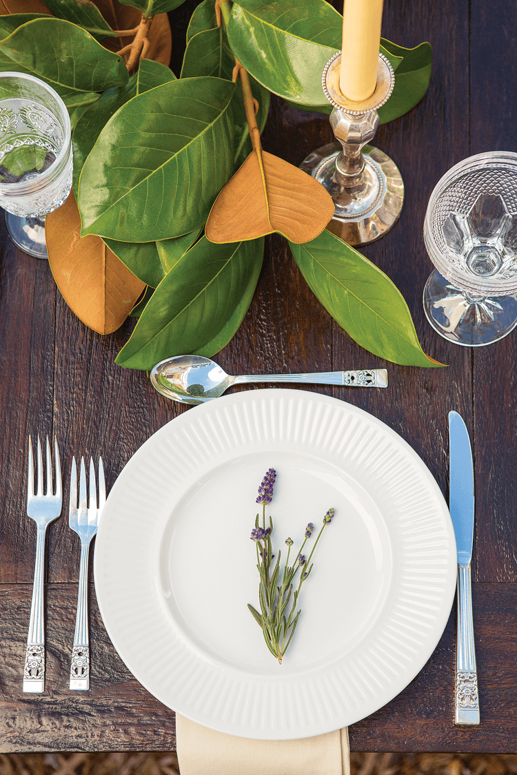 A simple table setting with silver flatware, white and crystal elements, and garnished with lavender and magnolia branches. 