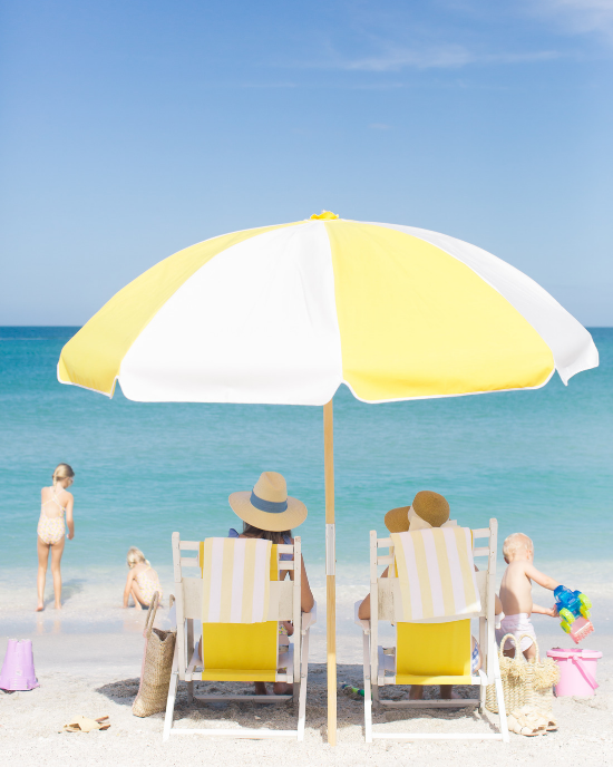 White and yellow beach umbrella shading two adults in beach chairs and surrounded by 3 small children playing in the sand.