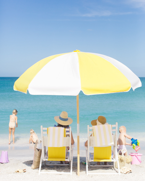 White and yellow beach umbrella shading two adults in beach chairs and surrounded by 3 small children playing in the sand. 