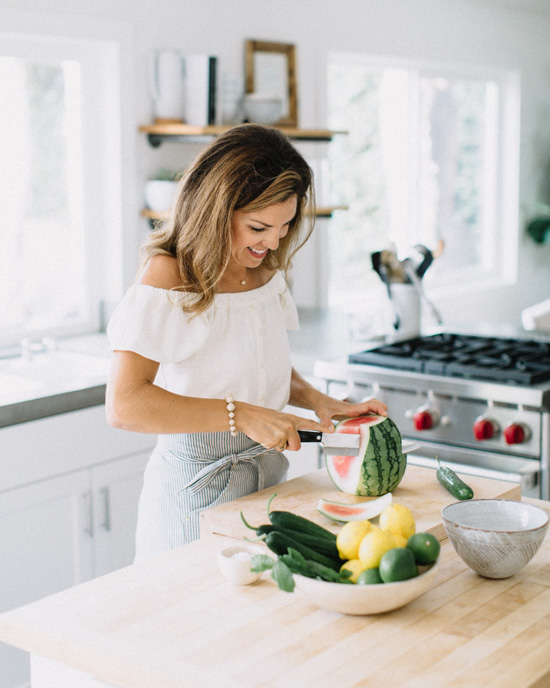 Chef, Sara Forte, standing at the butcher block island in her kitchen, cutting a watermelon. 