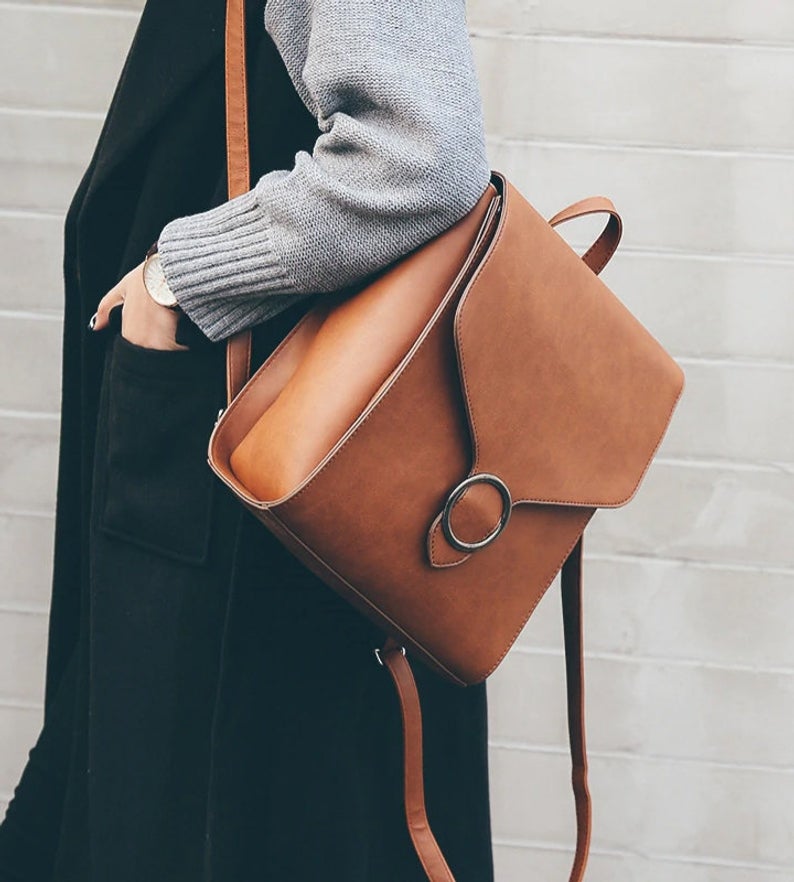 Woman in black jumper with chunky knit gray cardigan modeling a brown leather purse that also converts to a backpack. 