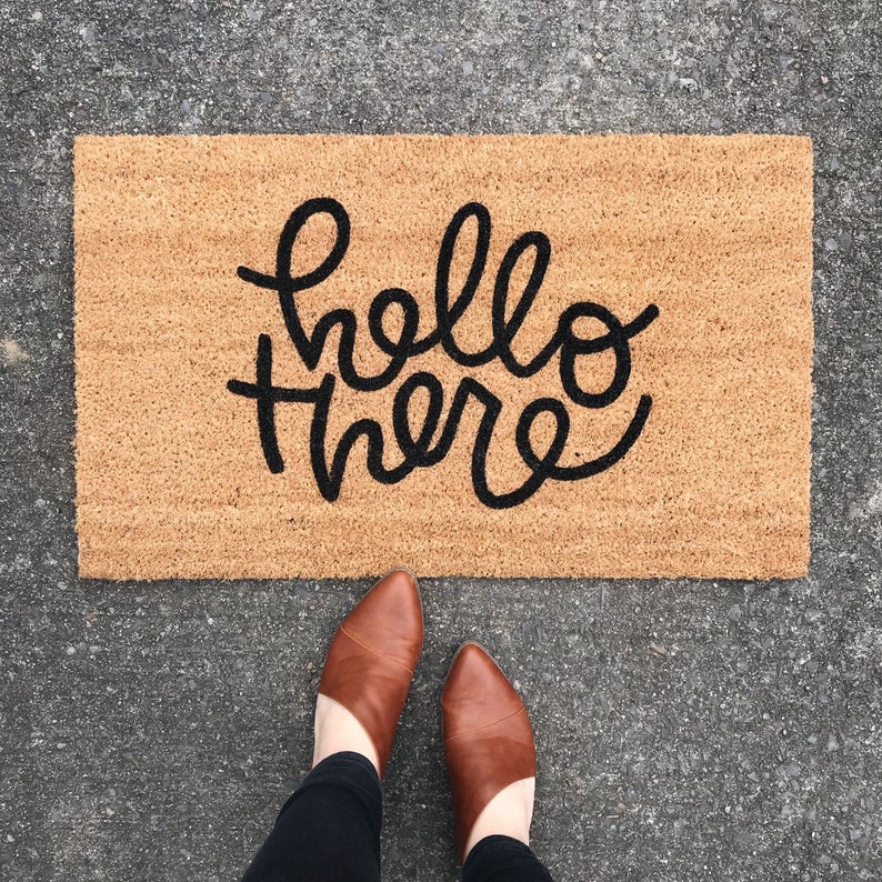 Jute doormat with "hello there" in black swirly font with feet in brown booties to show scale. 