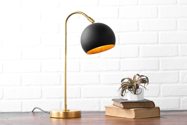 Wooden desk topped with an air plant on a stack of vintage books next to a black and brass dome lamp. 