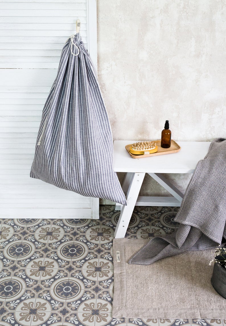 Cement tiled bathroom with a white bench and a hanging striped linen laundry bag hanging from a closet door.