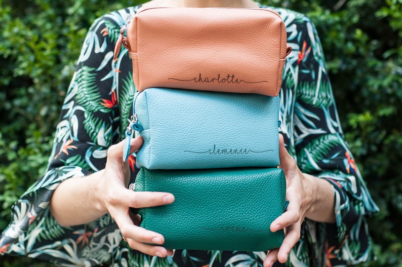 Girl in a tropical kimono holding a stack of leather, colored monogrammed makeup bags. 