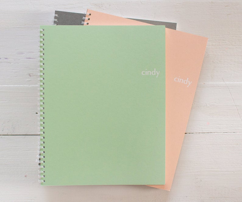 Stack of 3 spiral notebooks in gray, green and peach with monogrammed name on the cover.