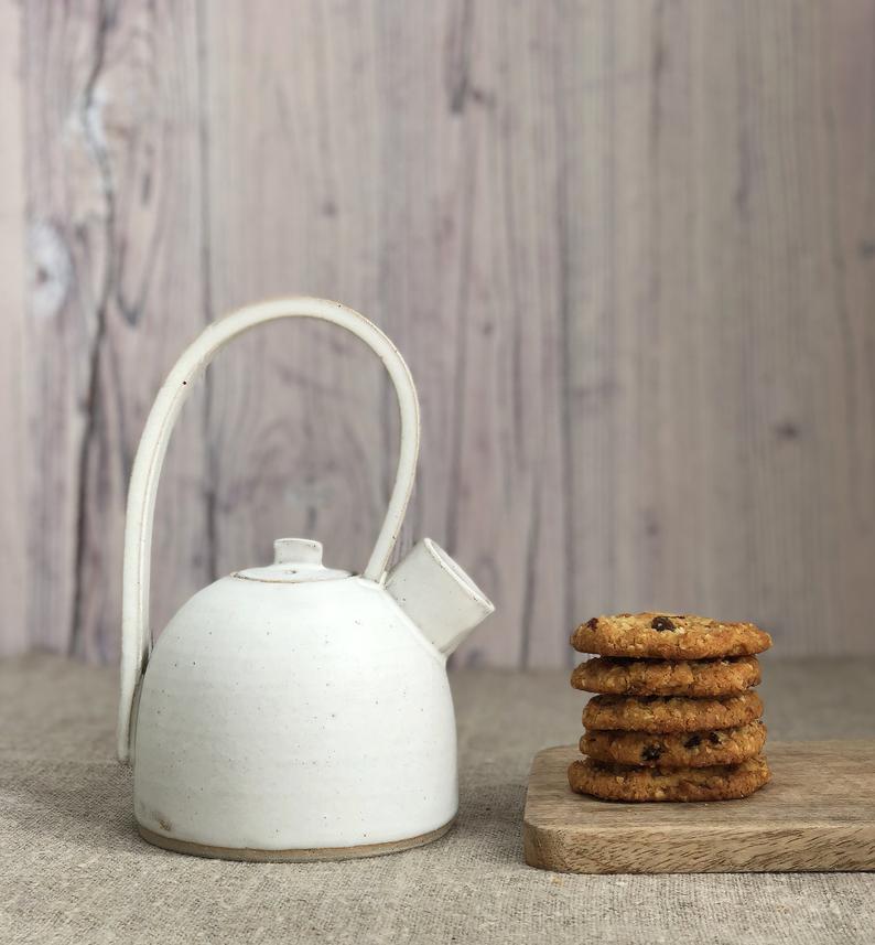 White, handmade ceramic teapot with tall handle places next to a pile of homemade cookies. 