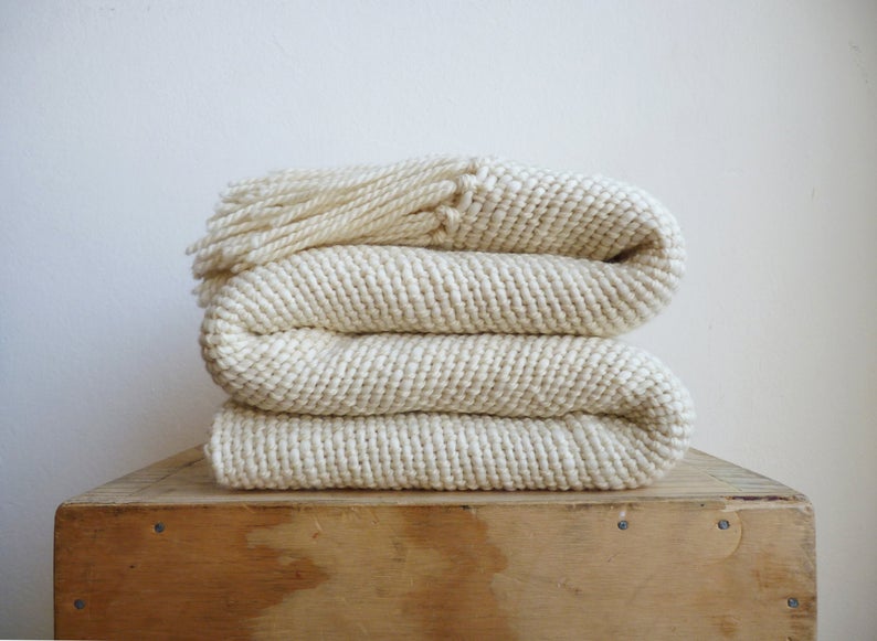 Cream colored handmade throw blanket with a fringed edge. Perfect for your dreamy dorm and folded compactly on top of a wooden box. 