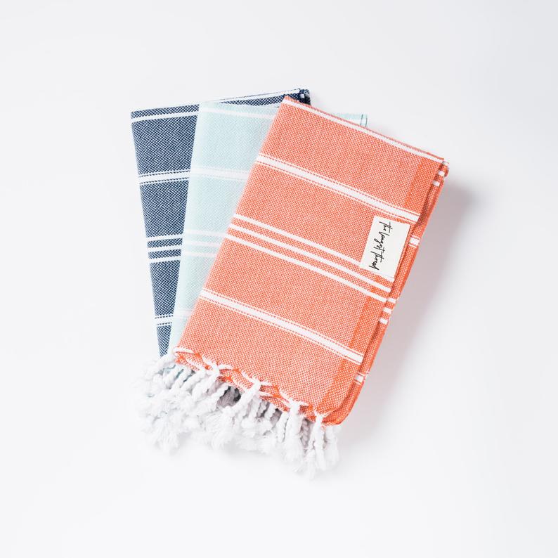 Stack of three Turkish towels in navy, light blue and coral all with white fringe. 