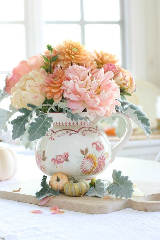 A hand-painted pitcher sitting on a wooden cutting board and filled with pink peonies and sherbet colored dahlias. 