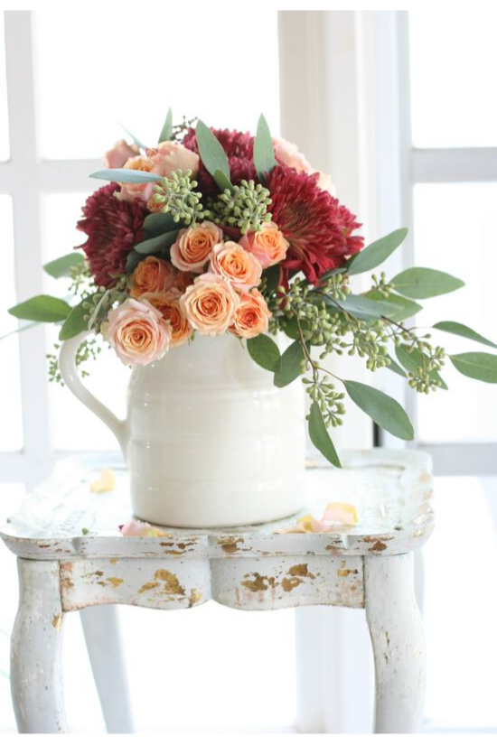 White pitcher filled with dahlias, roses and eucalyptus sitting on a distressed, white, wooden side table.