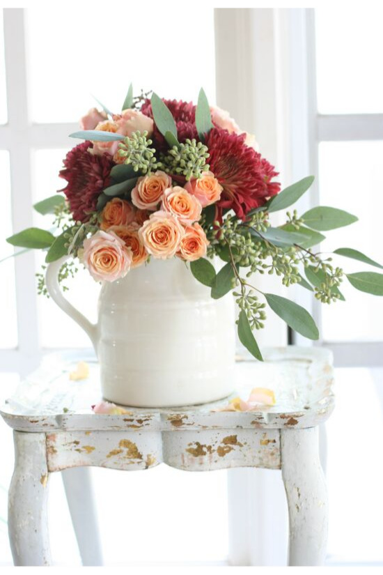 White pitcher filled with dahlias, roses and eucalyptus sitting on a distressed, white, wooden side table. 