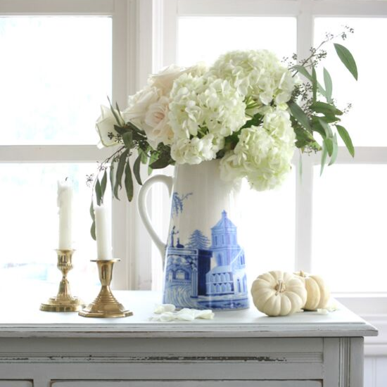 Light shades of hydrangeas, roses with eucalyptus accents in a white pitcher with cobalt blue accents posed next to brass candlesticks and small white pumpkins on a distressed sideboard in front of a window. 