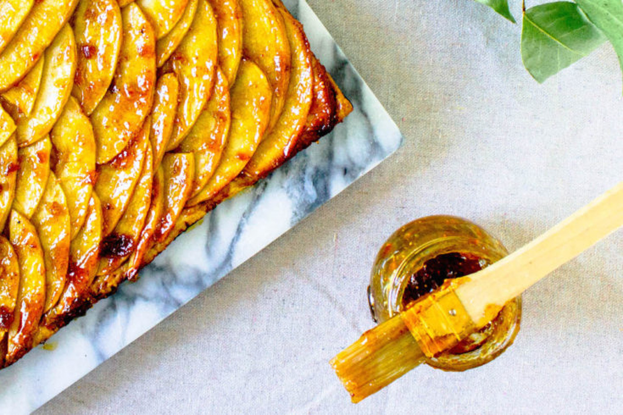 A marbled tray filled with fresh French Apple Tart pictured next to a jar topped with a gooey pastry brush.